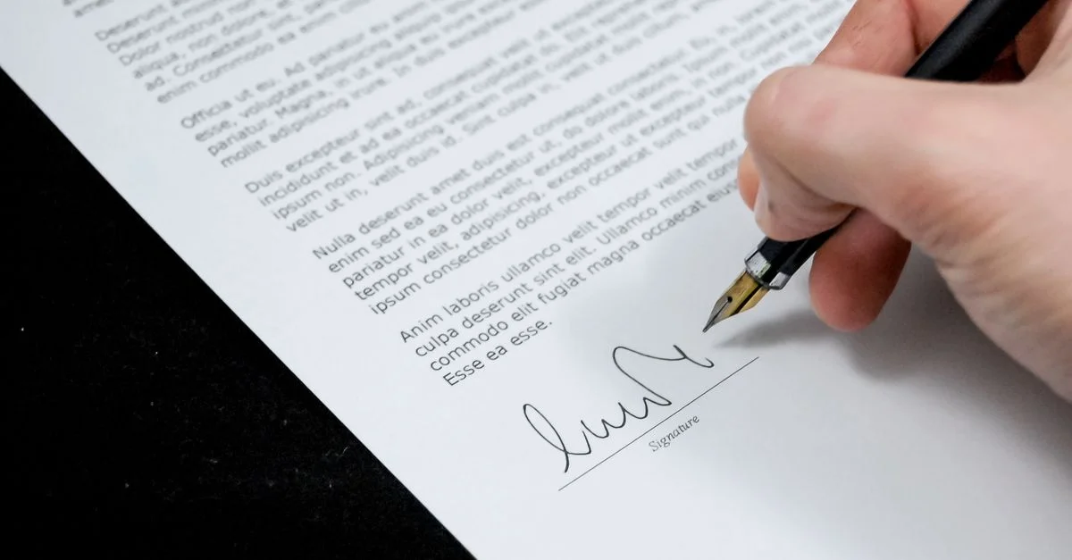 Primer plano de una mano firmando un documento formal con una pluma, indicando acuerdo.