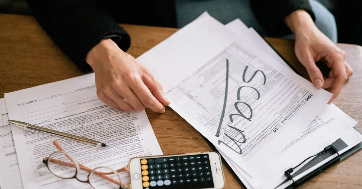 Manos de una persona examinando formularios de impuestos etiquetados como estafa, con calculadora y papeles.