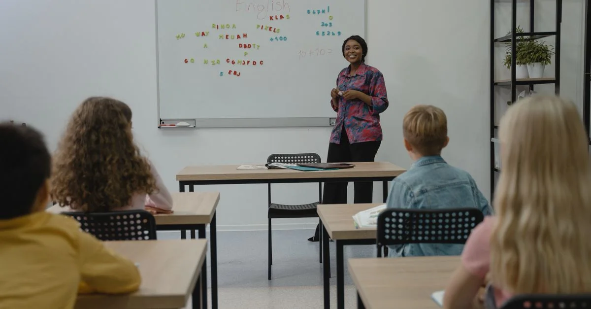 Un grupo diverso de estudiantes en una clase de inglés con una profesora sonriente frente a la pizarra.
