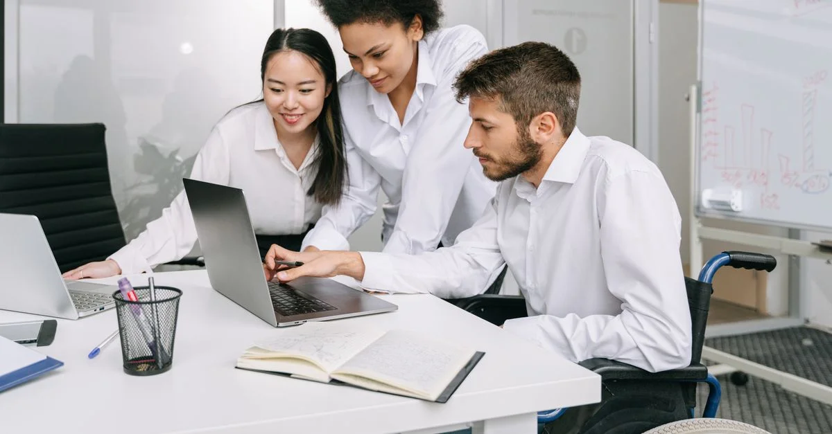 Compañeros de trabajo diversos en una oficina moderna colaboran en portátiles, mostrando trabajo en equipo e inclusión.