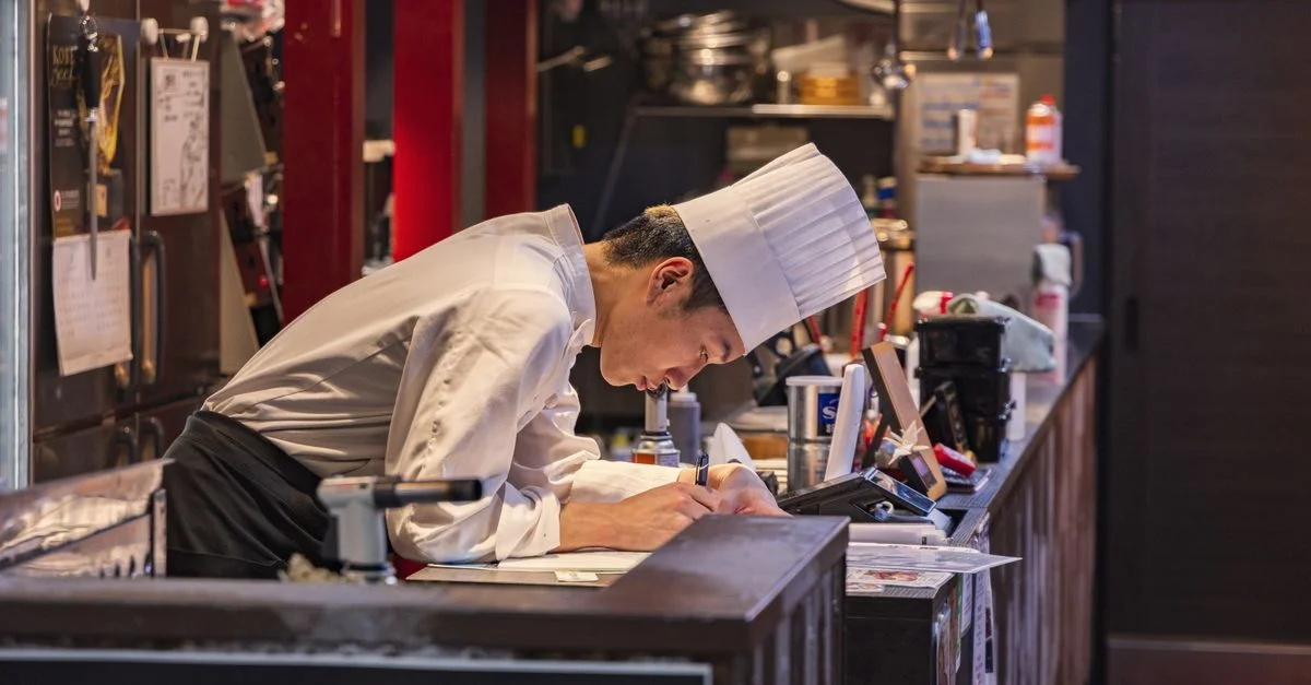 Chef en una cocina, enfocado en escribir con equipo culinario alrededor.