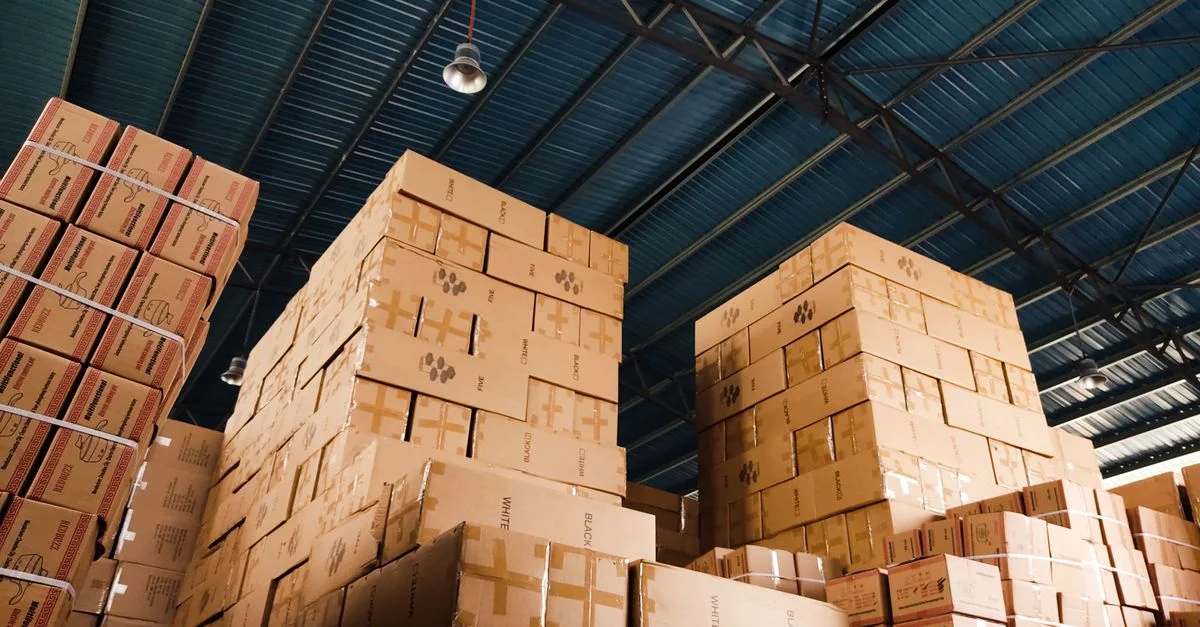 Altas pilas de cajas de cartón organizadas en un almacén con un techo de metal azul.