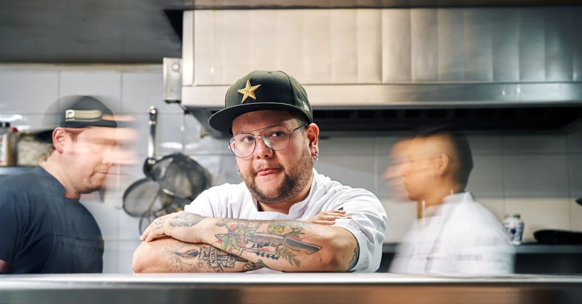 Chef confiado con tatuajes y gorra en una escena de cocina bulliciosa, destacando el arte culinario.