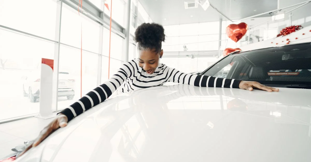 Mujer afroamericana abrazando con alegría un coche en un concesionario con globos en forma de corazón.
