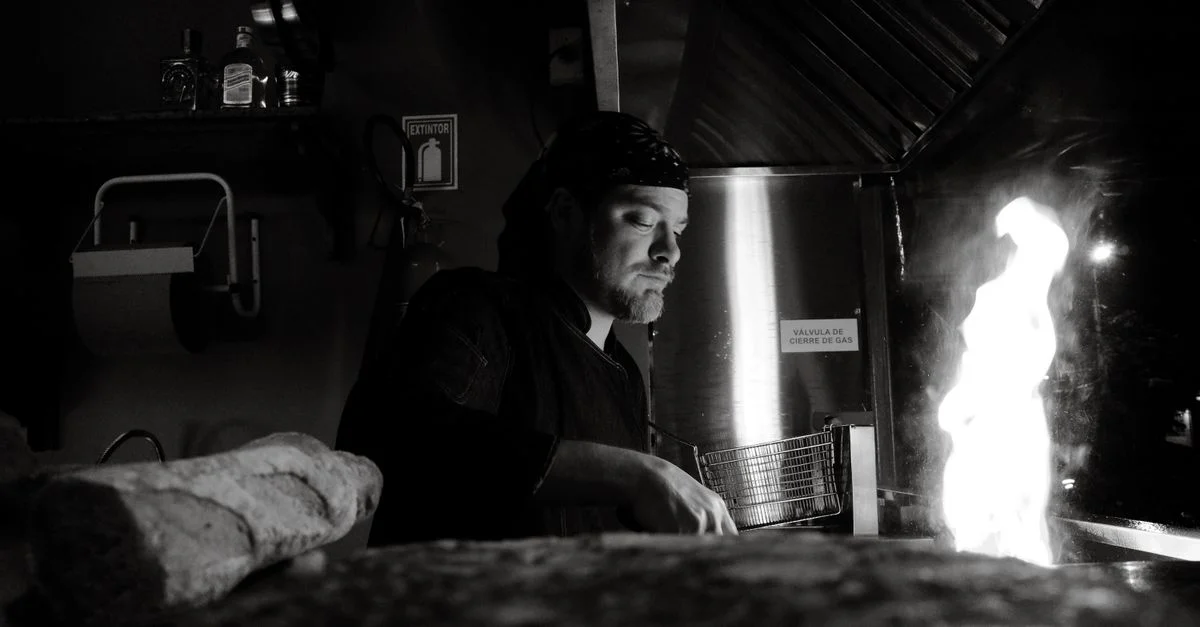 Un chef prepara hábilmente comida sobre una llama abierta en una cocina comercial, en blanco y negro.
