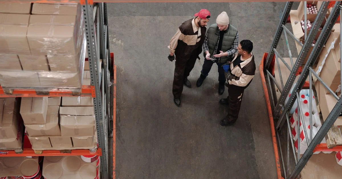 Tres trabajadores de almacén discutiendo logística en un área de almacenamiento industrial.