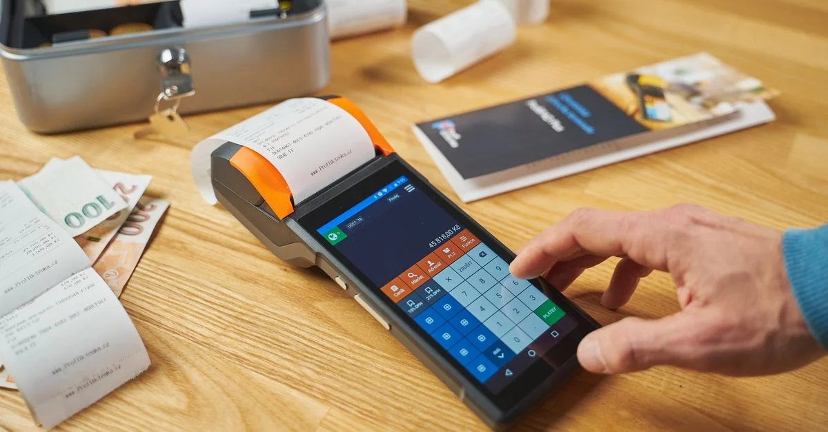 Persona utilizando un terminal de pago digital en un escritorio de madera con dinero y recibos visibles.