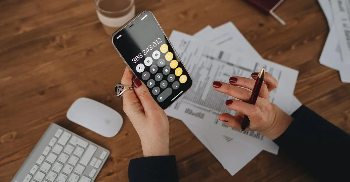 Mujer calculando gastos utilizando una calculadora de smartphone en un escritorio de madera con documentos.