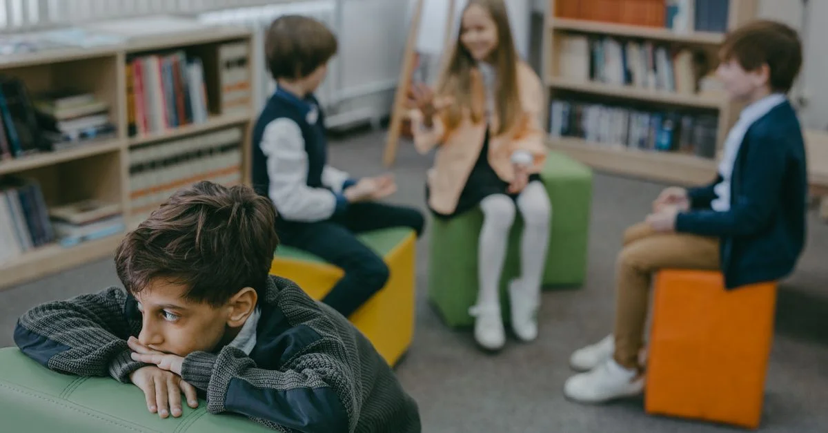 Un niño triste aislado en una biblioteca escolar mientras sus compañeros charla en el fondo.