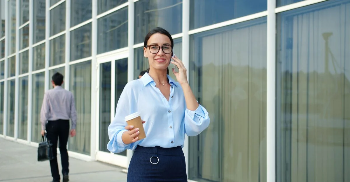 Una mujer sonriente, con una taza de café en la mano, habla por teléfono frente a un moderno edificio de oficinas.