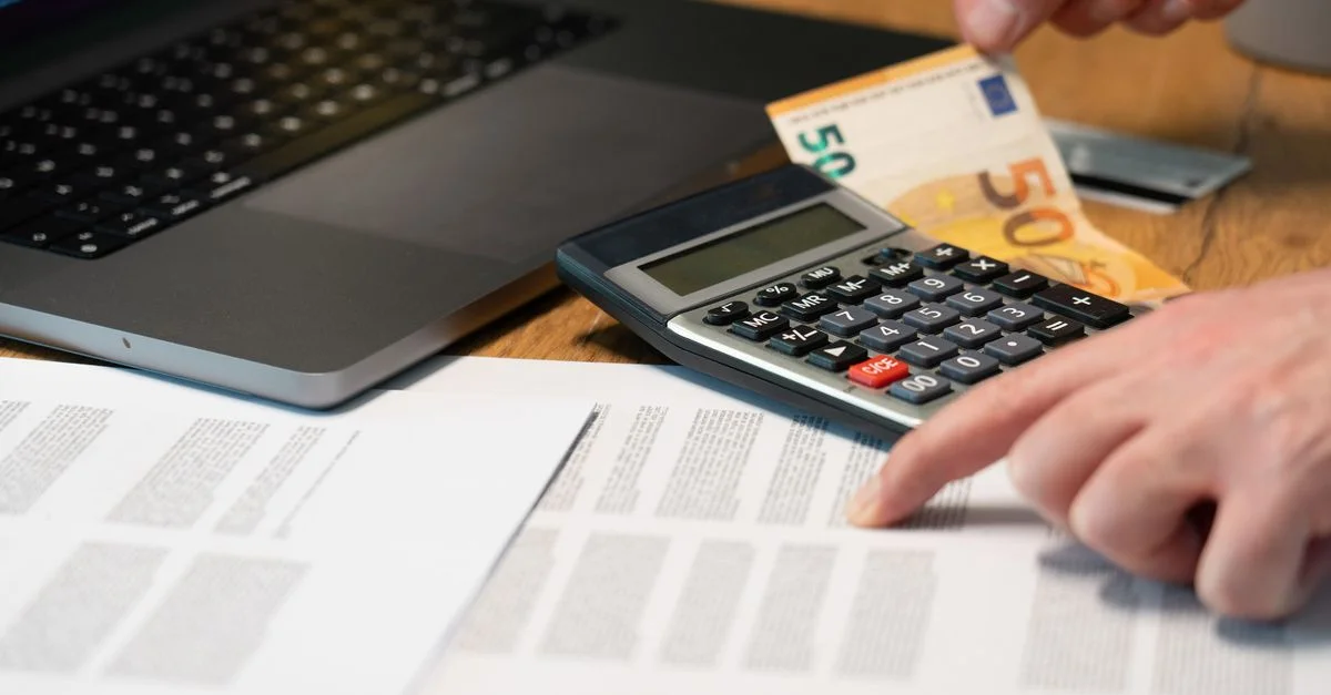 Primer plano de manos usando calculadora con billetes de euro y portátil en la mesa.