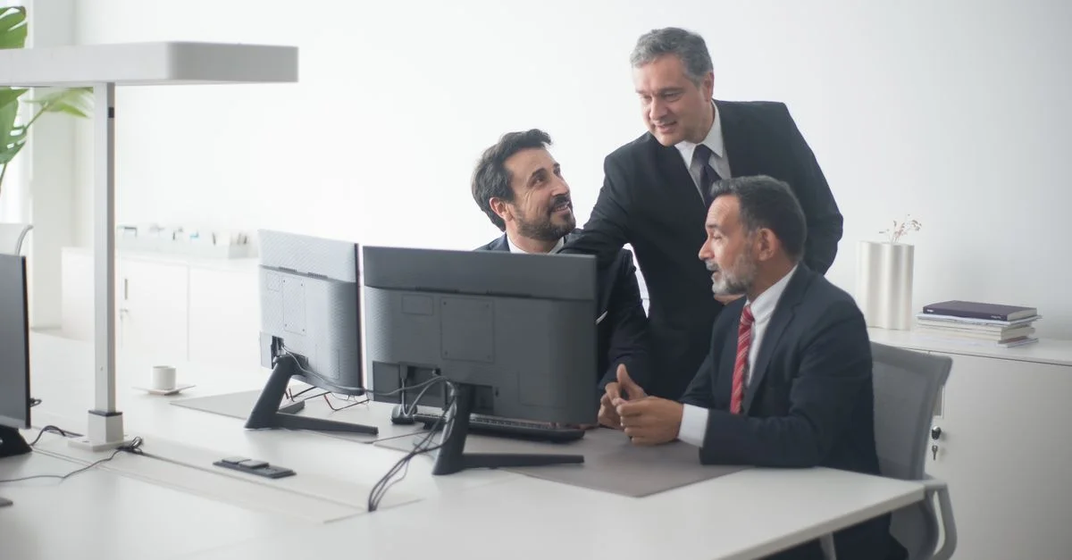Tres ejecutivos de negocios con trajes colaborando alrededor de monitores dobles en un entorno de oficina moderna.