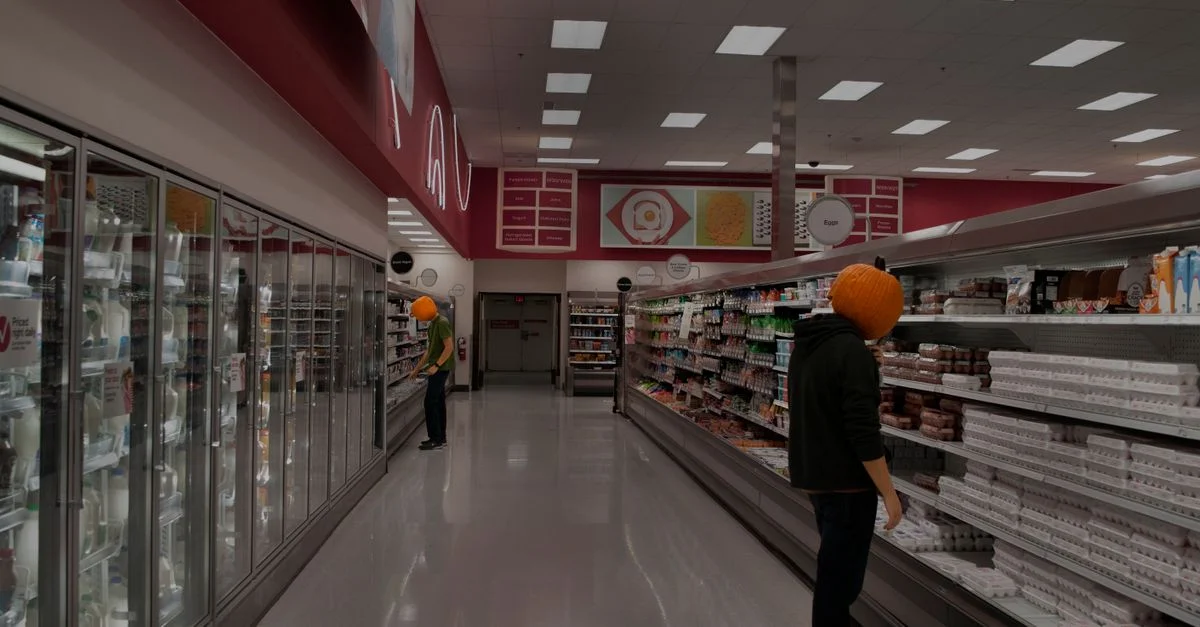 Dos personas con máscaras de calabaza recorriendo un pasillo de supermercado lleno de comestibles.