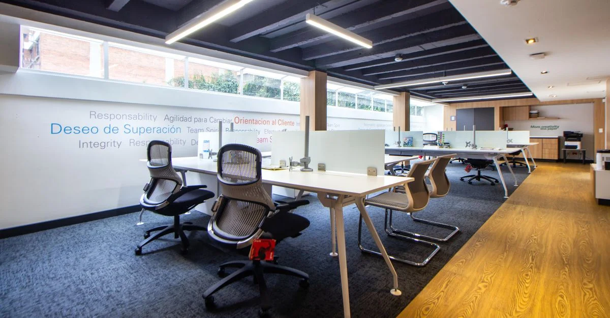 Un espacio de oficina moderno y bien iluminado con sillas ergonómicas, mesas y divisores de vidrio.