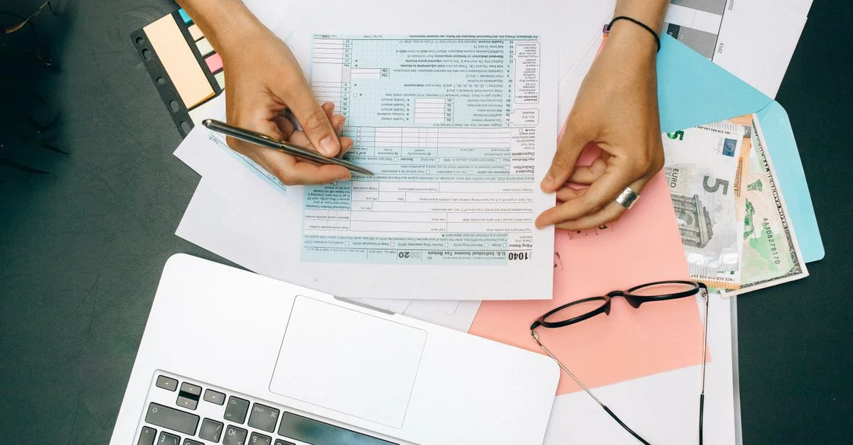 Manos escribiendo en documentos fiscales con laptop, gafas y dinero en la mesa.