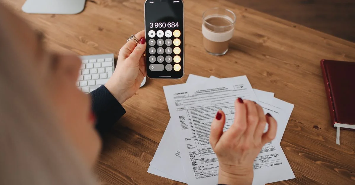 Una mujer calcula en su smartphone mientras trabaja con documentos en un escritorio.