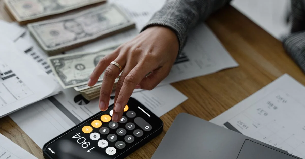 Primer plano de una persona usando una calculadora de smartphone entre dinero y documentos financieros en una mesa de madera.