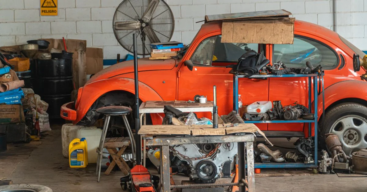 Coche clásico rojo siendo reparado en un garaje desordenado, lleno de herramientas y piezas de automóviles.