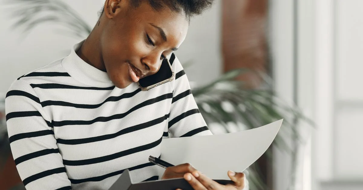 Joven mujer africana multitarea en una llamada telefónica y tomando notas en interiores, vestida con rayas.