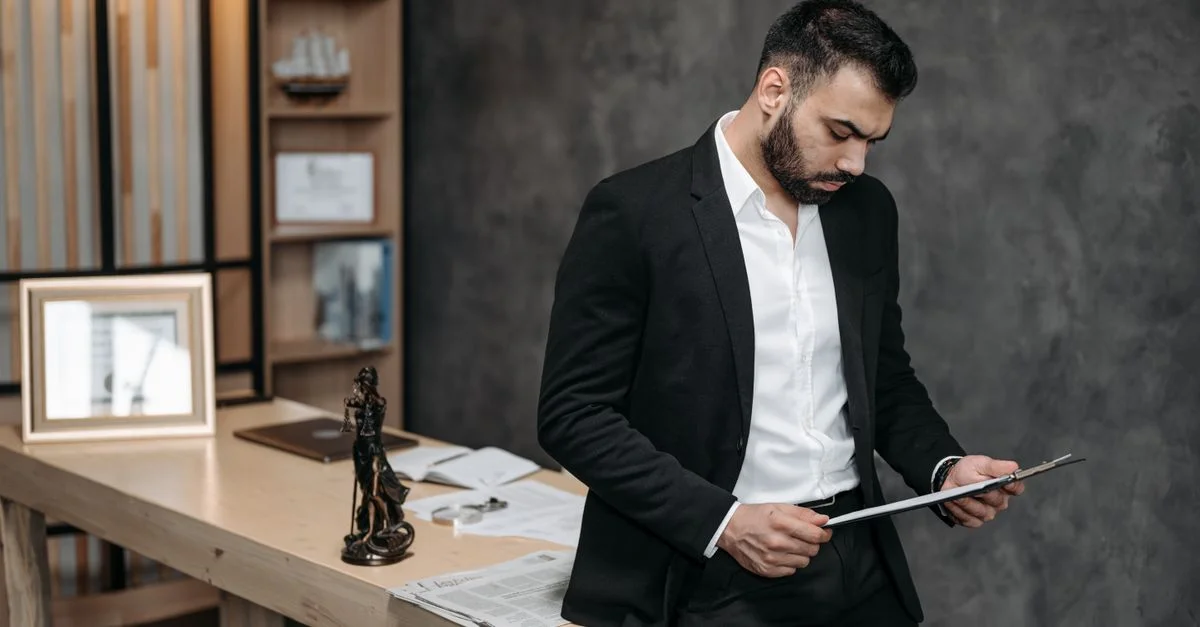 Un abogado masculino con traje negro concentrado en documentos en un entorno de oficina.