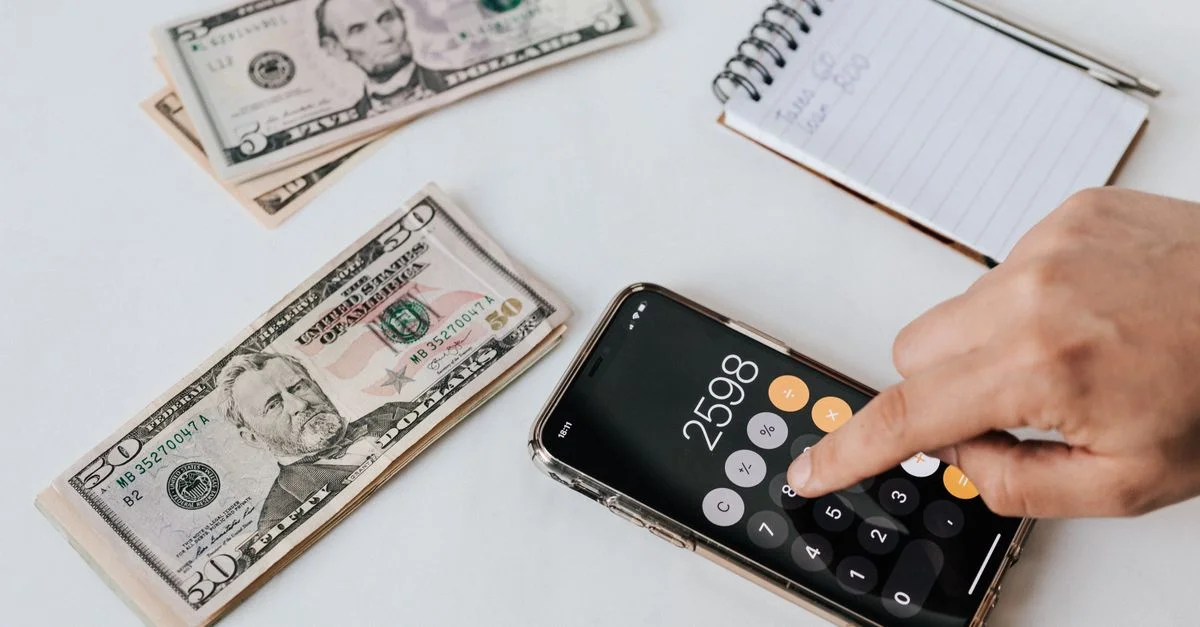 Mano usando una calculadora de smartphone con billetes de dólar estadounidense y una libreta para planificación financiera.