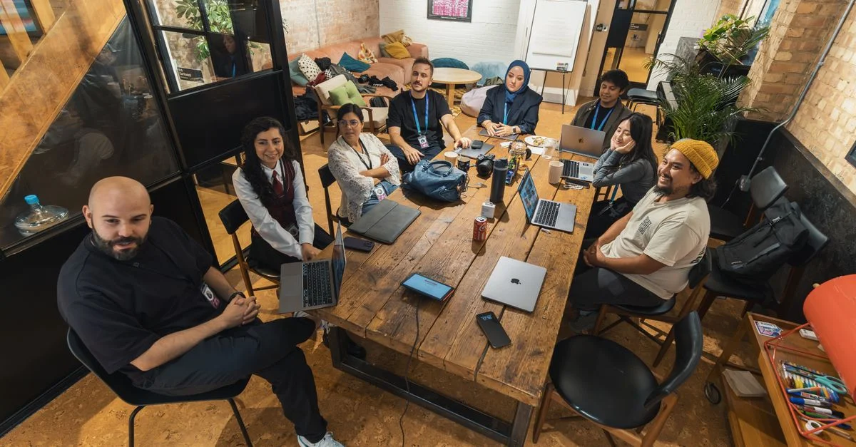 Grupo de colegas reunidos en torno a una mesa en un entorno de oficina moderna, colaborando en laptops.