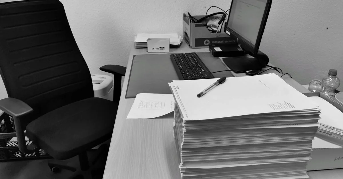 Imagen en blanco y negro de un escritorio de oficina desordenado con un ordenador, silla y documentación.