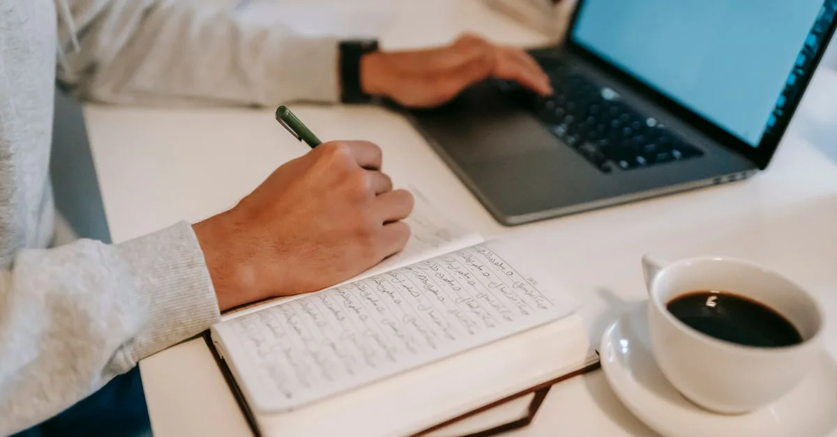 Un primer plano de una persona escribiendo en un cuaderno mientras usa un laptop, acompañada de una taza de café.