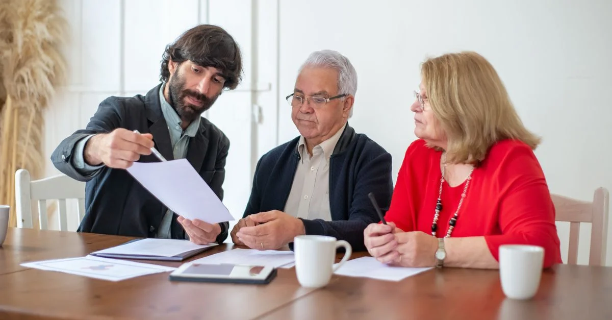 Pareja de ancianos discutiendo documentos con un asesor en una mesa de madera en interiores.