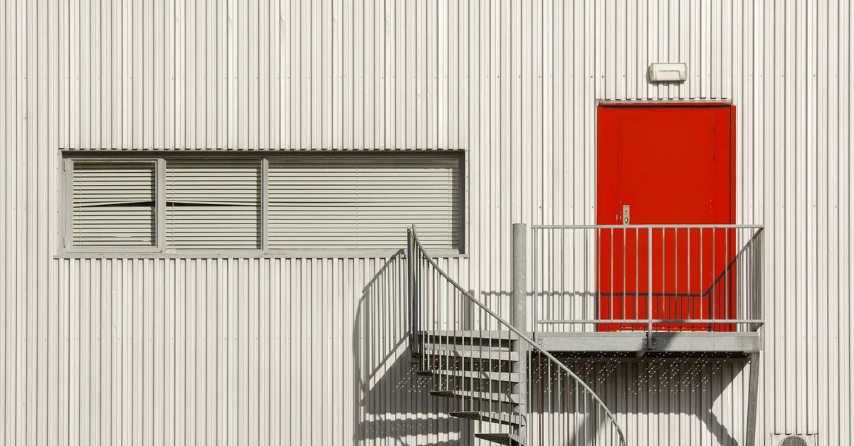 Puerta roja audaz en un edificio industrial blanco con escalera de caracol.