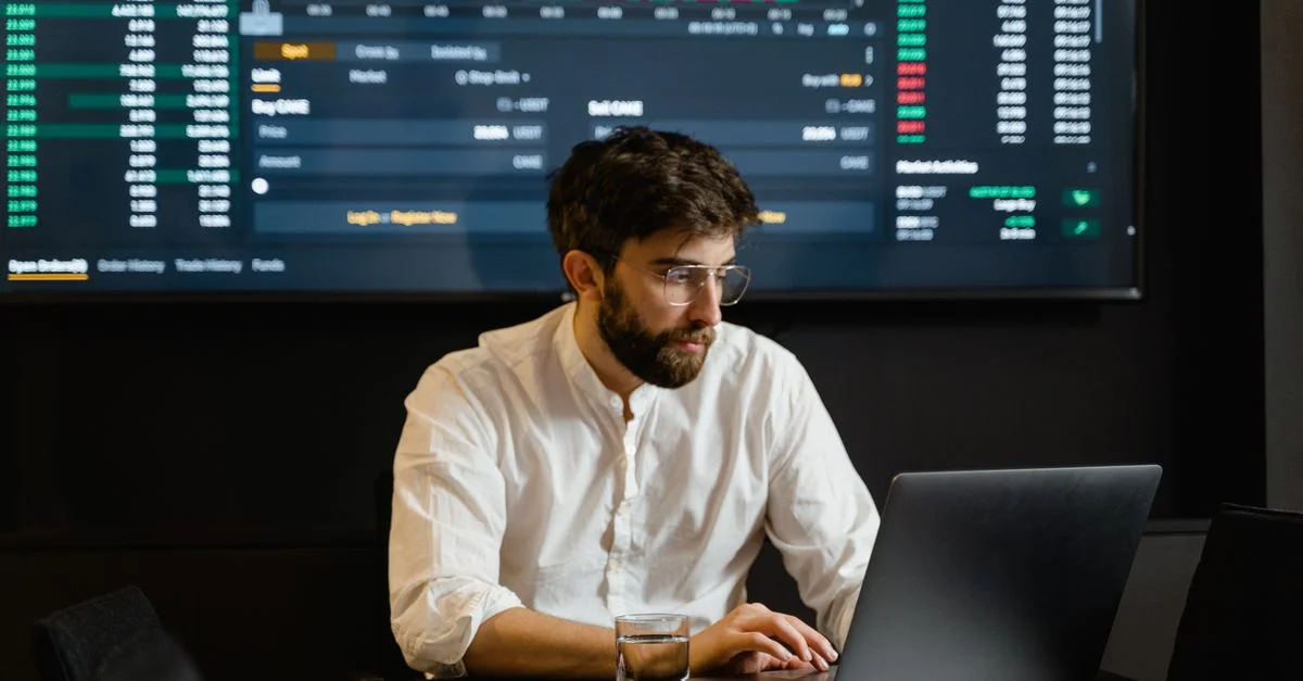 Profesional analizando tendencias del mercado de valores usando laptop y pantalla digital en una oficina moderna.