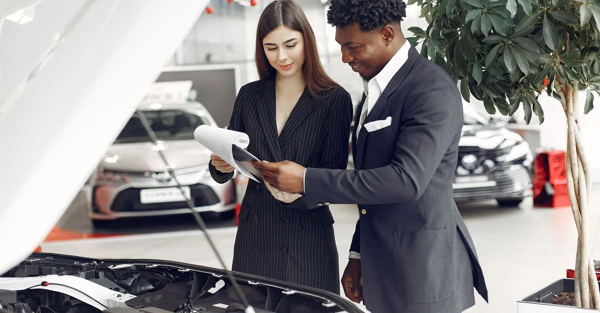 Hombre y mujer revisando documentos en una concesionaria de coches junto a un capó de coche abierto.