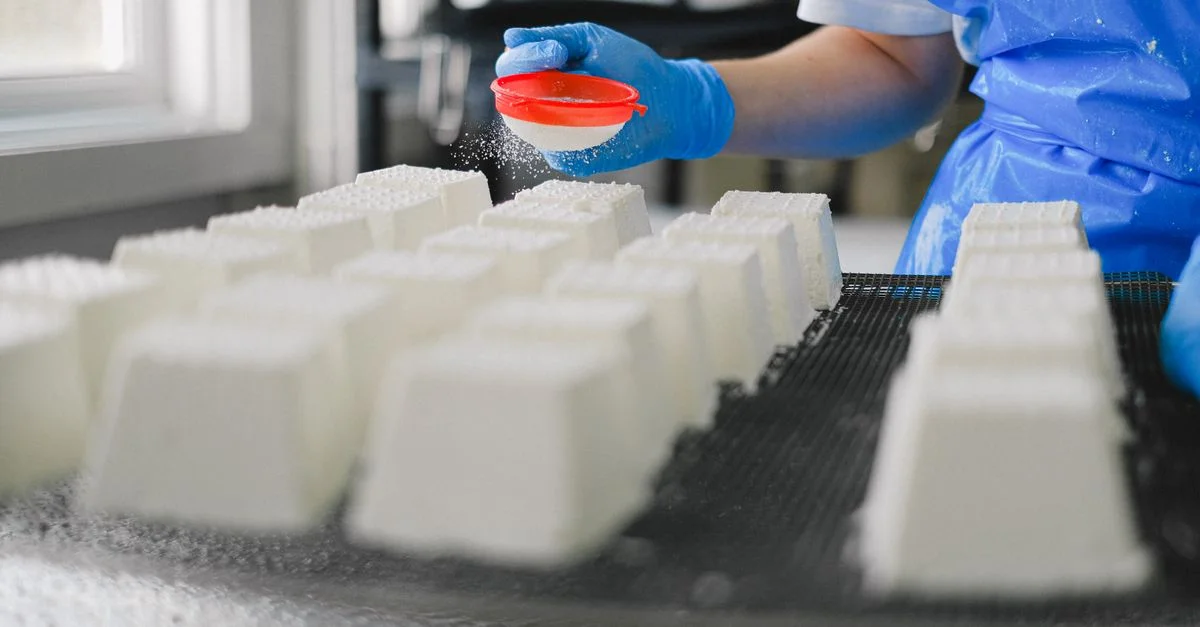 Close-up de un trabajador con guantes, espolvoreando queso en un entorno de fábrica, mostrando precisión.