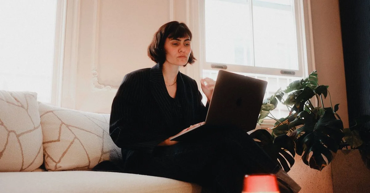Mujer en traje usando una computadora portátil en un sofá en una habitación luminosa, estilo de vida moderno.