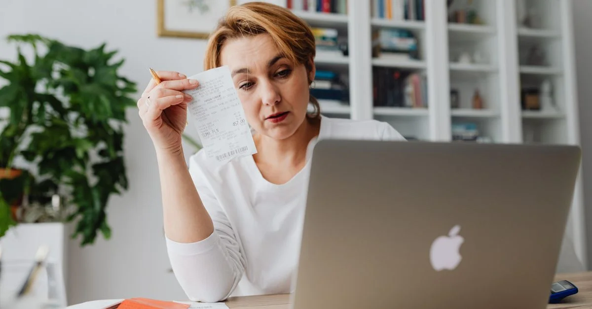 Mujer concentrada en recibos mientras usa un portátil, reflejando estrés financiero.