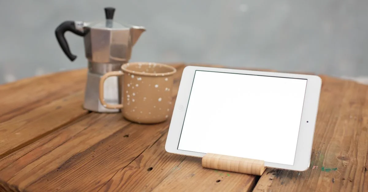 Mockup de una tablet junto a una cafetera y una taza sobre una mesa rústica.