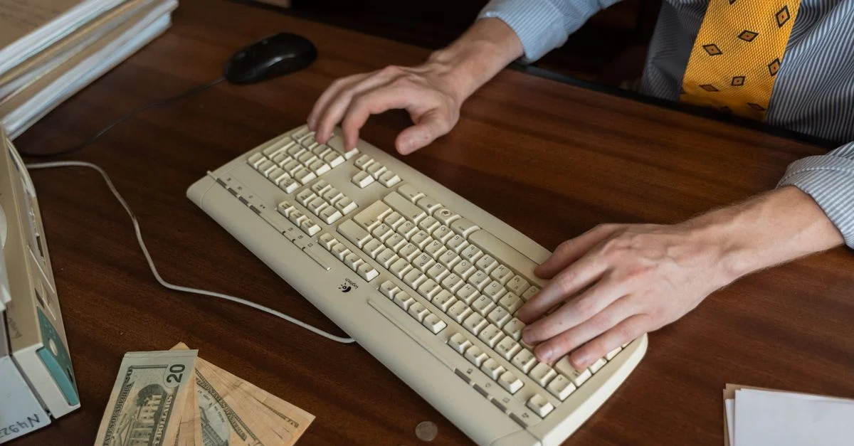 Manos escribiendo en un teclado vintage sobre un escritorio de madera con dinero y monedas.