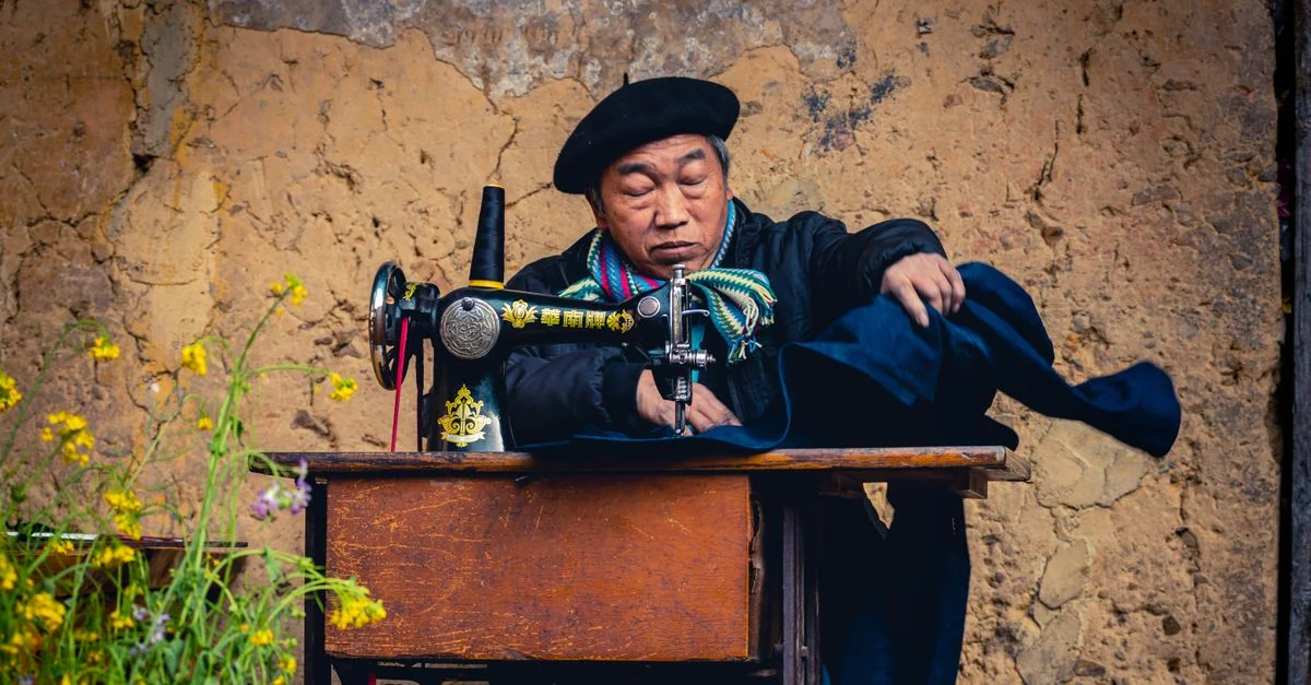 Un hombre adulto asiático cosiendo al aire libre en una máquina de coser vintage, vistiendo una bufanda y una boina coloridas.