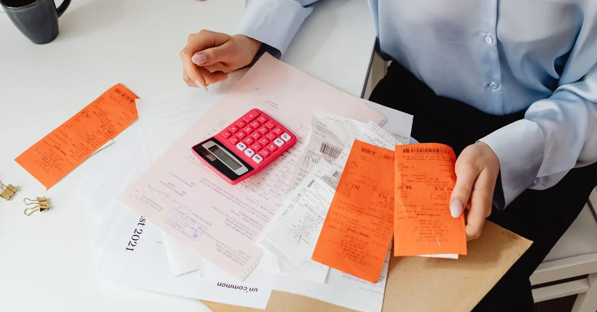 Una mujer revisa recibos y calcula gastos en un escritorio con una calculadora rosa.