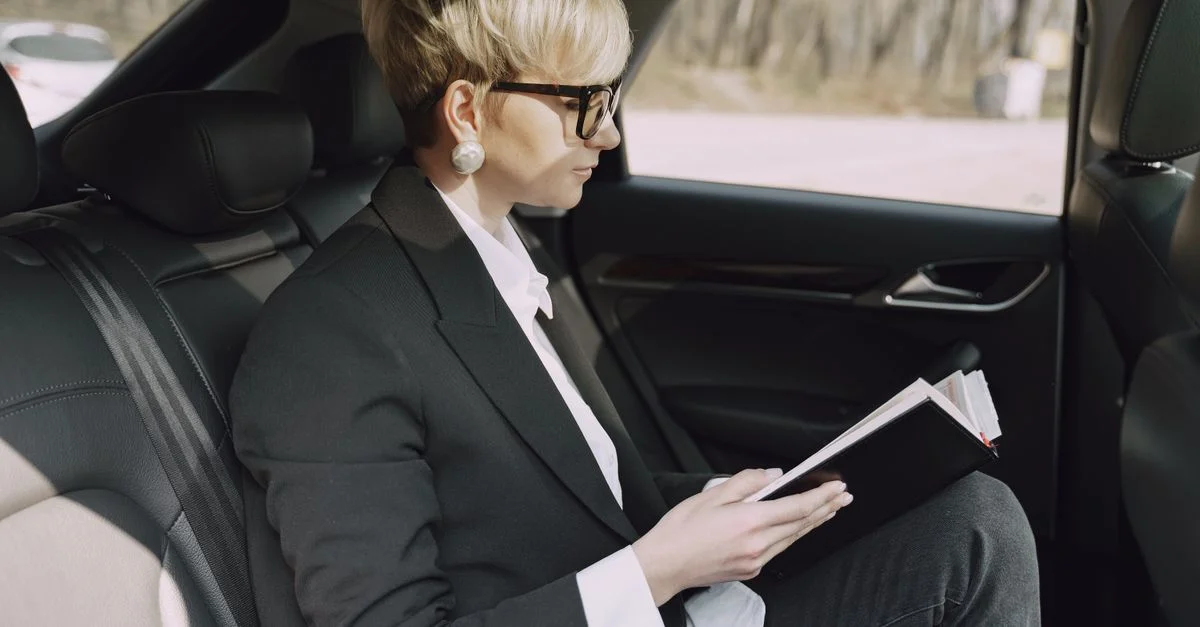 Mujer de negocios elegante concentrada en leer un cuaderno mientras está sentada en el asiento trasero de un coche durante el día.