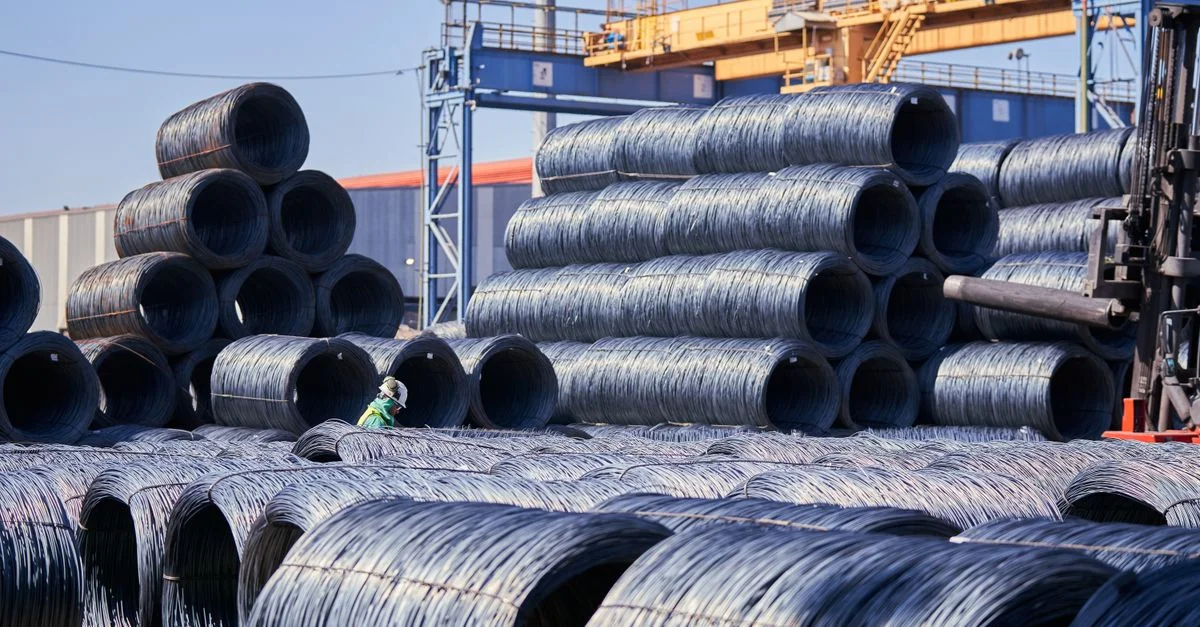 Grandes pilas de bobinas de alambre de acero almacenadas al aire libre en un sitio industrial bajo una brillante luz diurna.