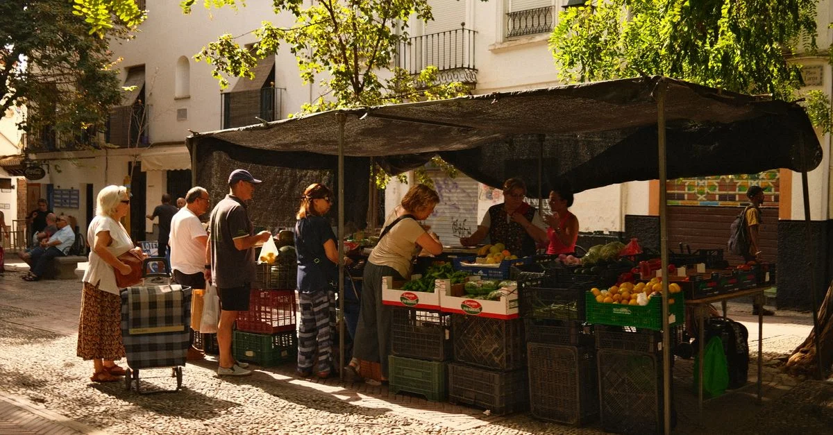 Un bullicioso mercado callejero con personas comprando productos frescos en un entorno urbano soleado.
