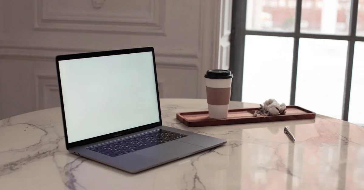 Un ambiente minimalista que presenta un portátil, una taza de café y un bolígrafo sobre una mesa de mármol junto a una ventana.