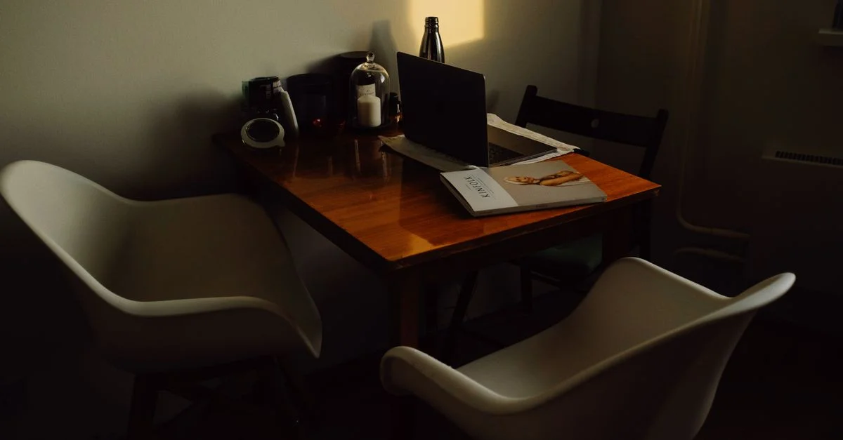 Una oficina en casa cálida y de tonos oscuros con una mesa de madera y sillas contemporáneas. Incluye un portátil, revista y decoración.