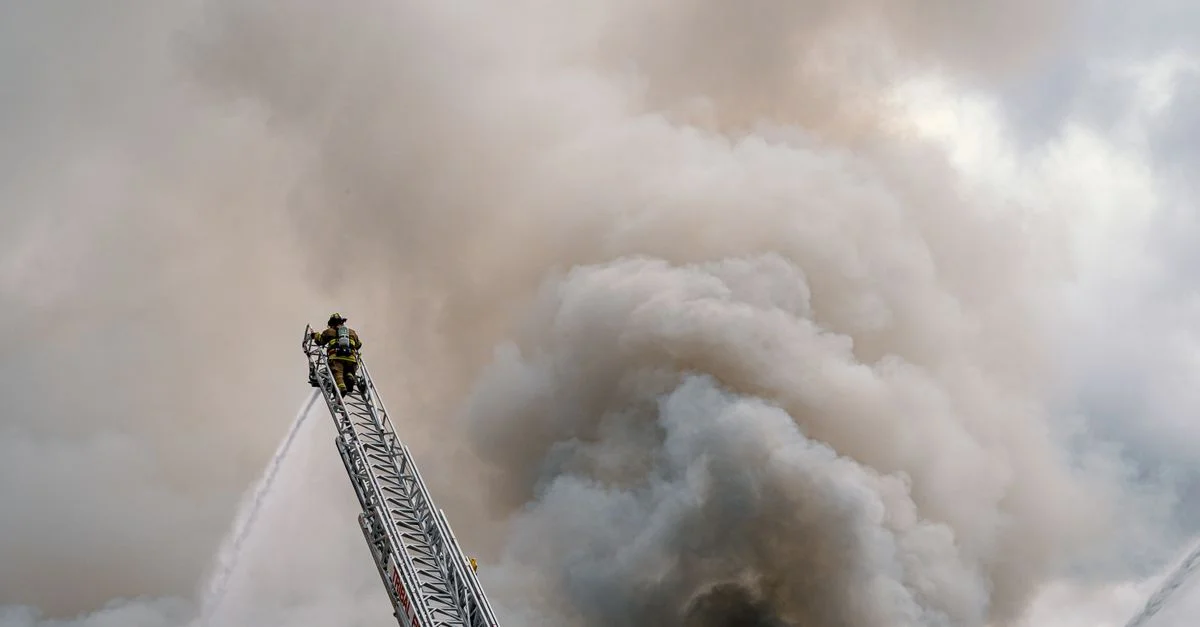 Un valiente bombero combate un gran incendio desde una escalera alta, envuelto en espesa humo.