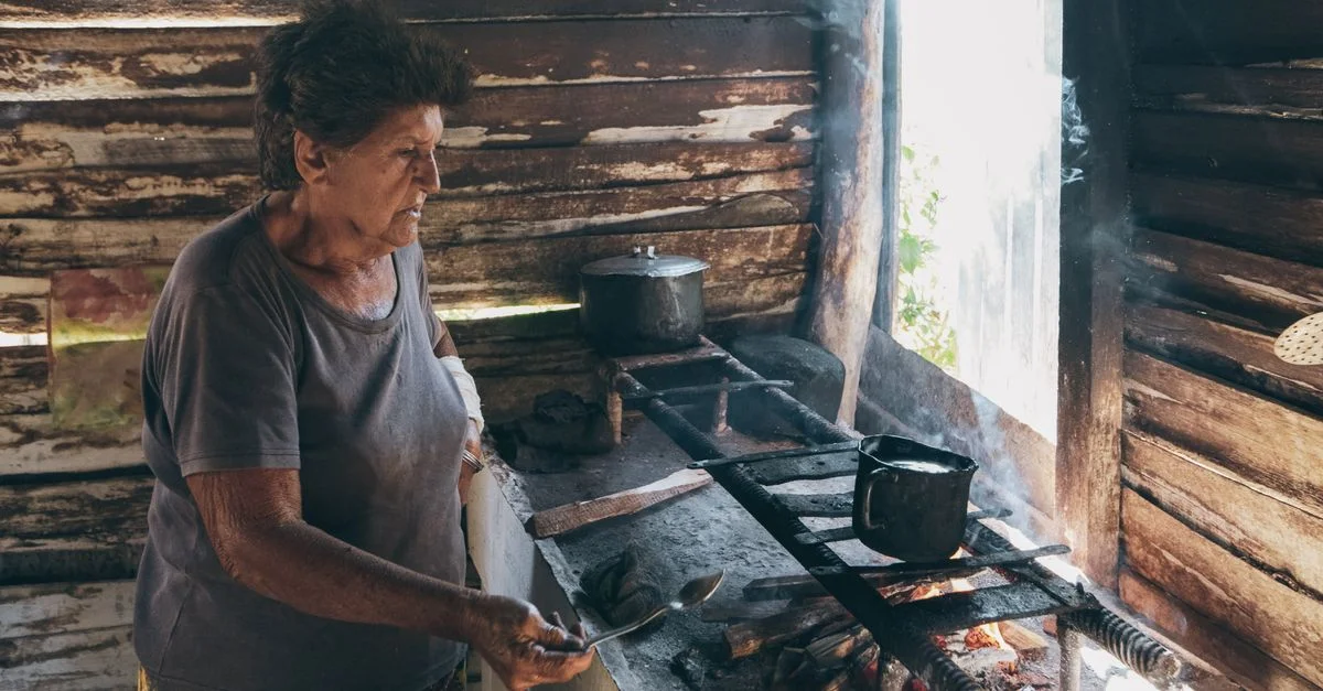 Mujer mayor preparando comida en un fuego abierto en una cabaña rústica de madera, rodeada de humo y calor.