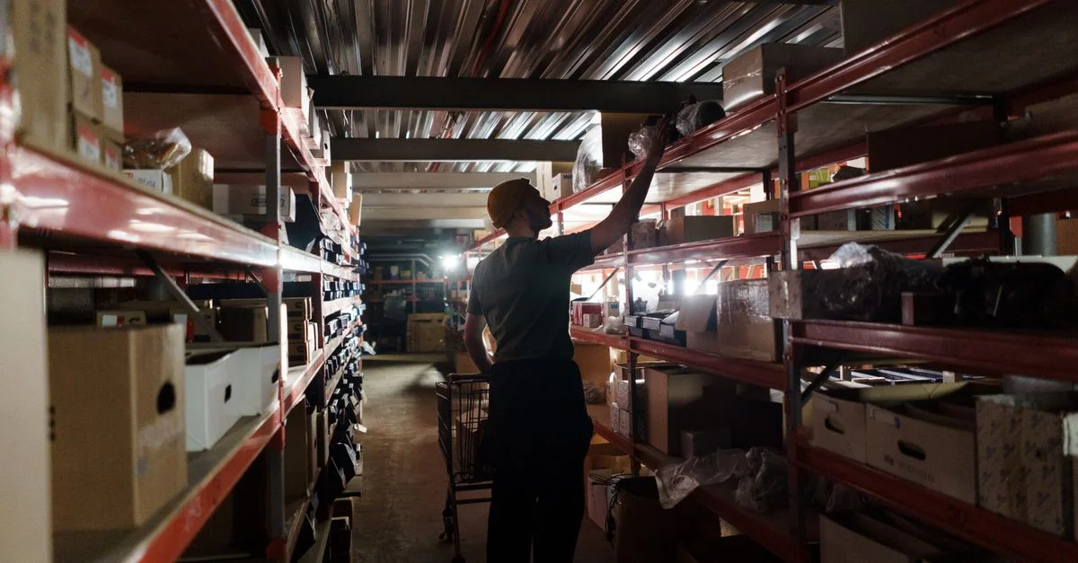 Un trabajador organizando el inventario en estanterías en un almacén poco iluminado, utilizando prácticas de almacenamiento eficientes.