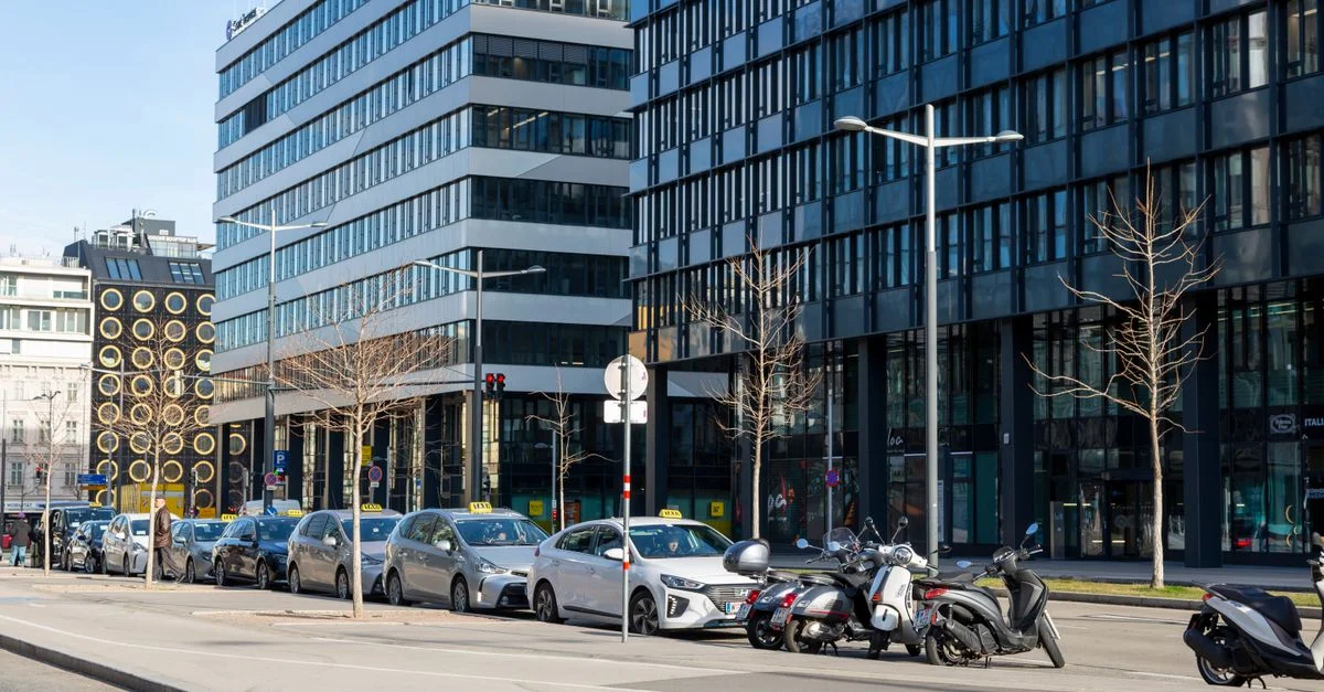 Calle urbana contemporánea con coches y motos aparcados junto a modernos edificios de oficinas.