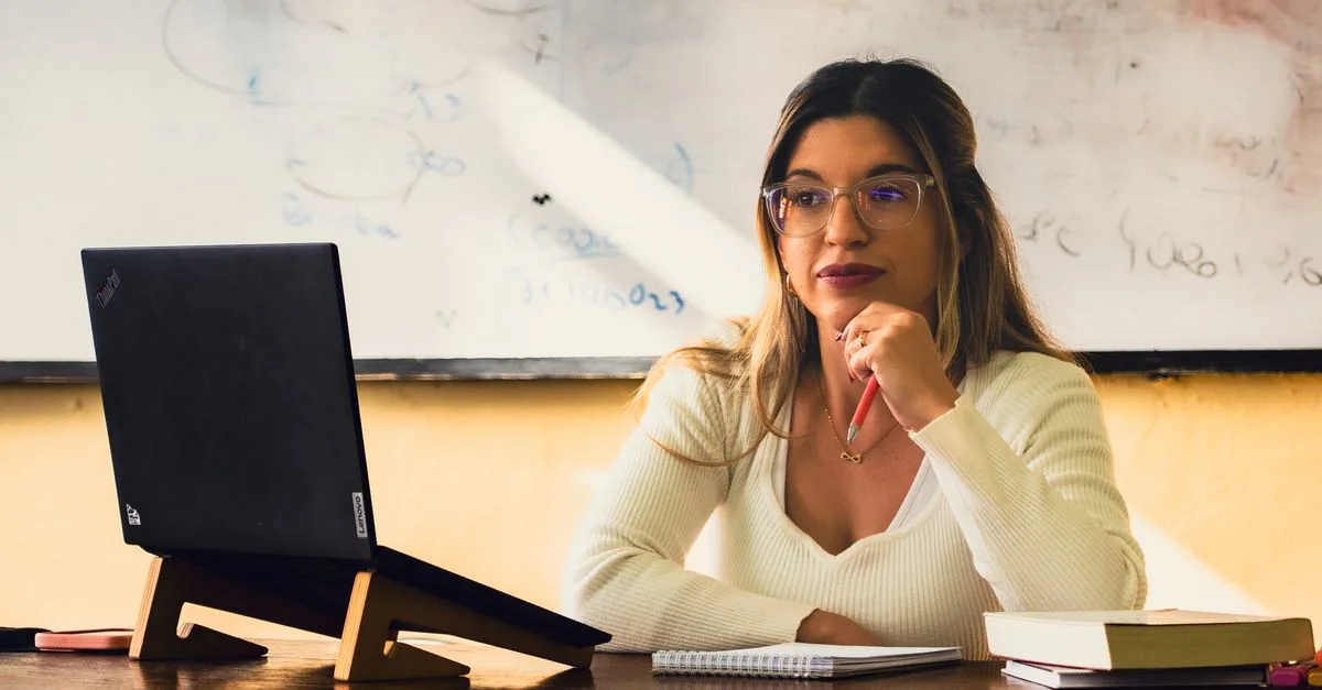 Mujer joven en un entorno de oficina trabajando en su computadora portátil, con expresión pensativa, rodeada de libros y apuntes.
