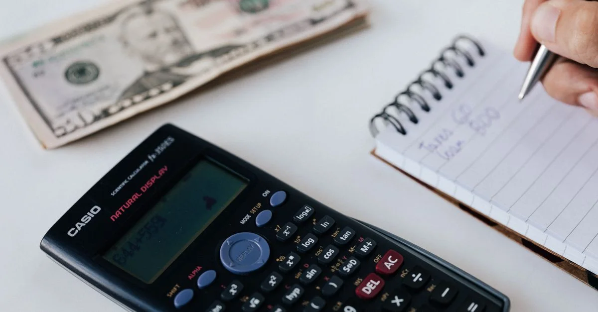 Acercamiento de una calculadora, billete de dólares estadounidenses y una mano escribiendo en un cuaderno, simbolizando la planificación financiera.